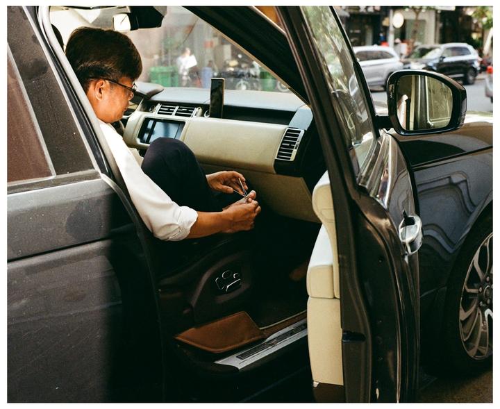 A man wearing glasses and a white shirt is sitting in the driver's seat of a car with the door open, trimming his fingernails.