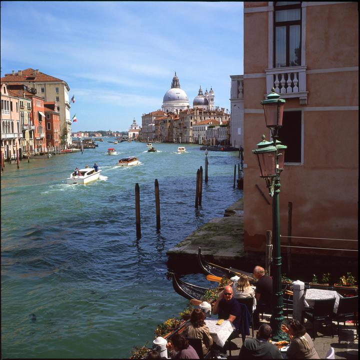 A group of people are seated at an outdoor cafe next to a canal with boats and historic buildings in the background.