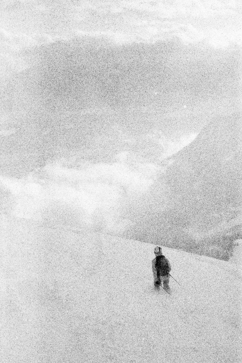 A skier stands on a snowy slope with mountains and clouds in the background.