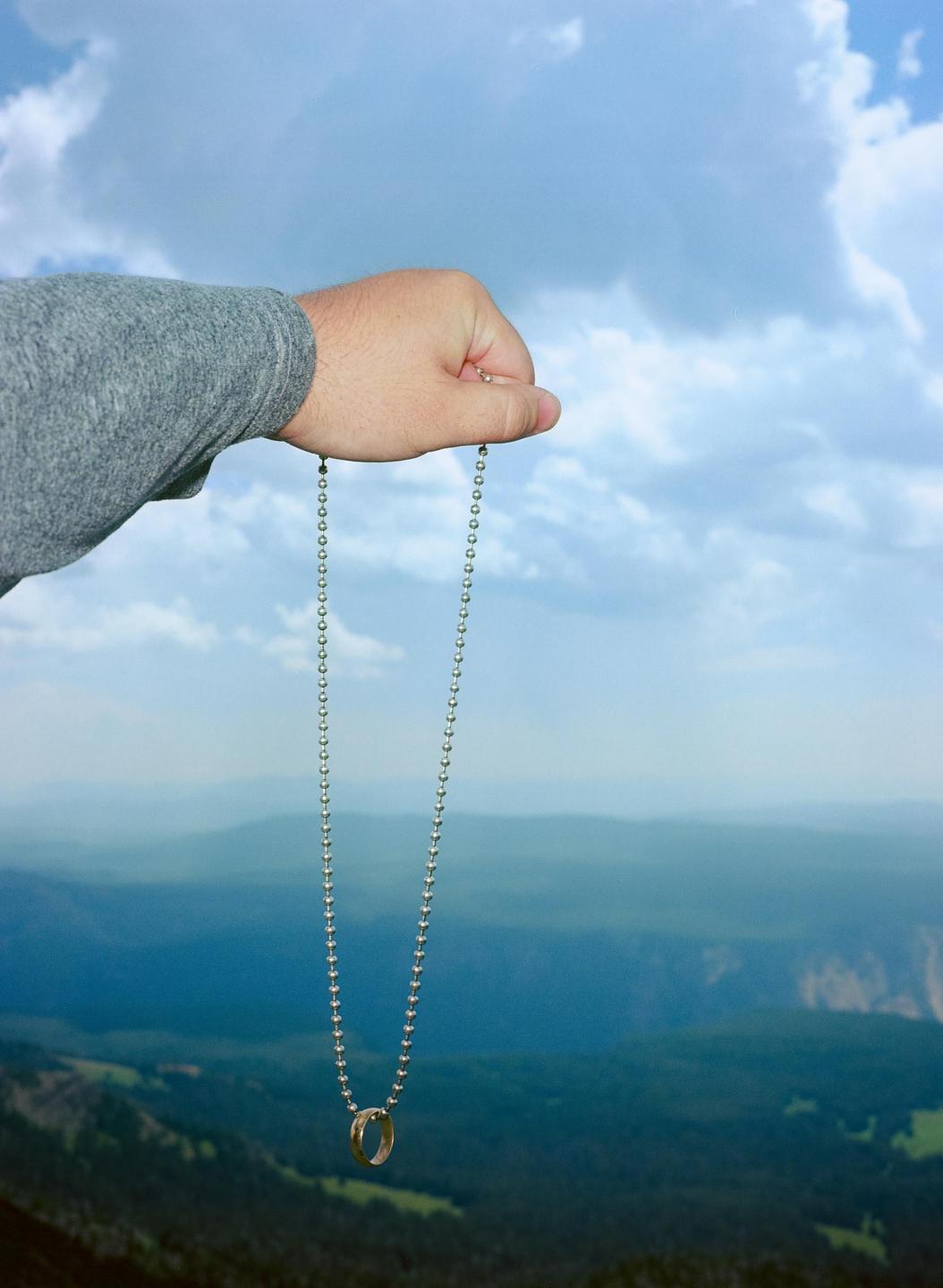 A man's arm extends into the frame, holding a chain with a ring at the end against a mountainous landscape under a cloudy sky.