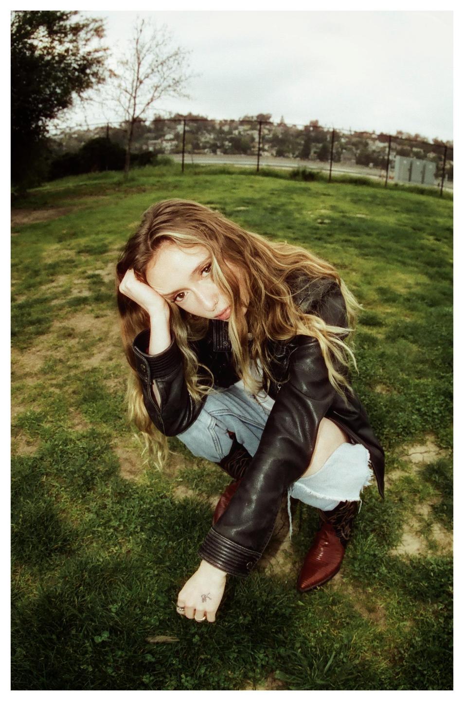 A woman with long hair, wearing a leather jacket, ripped jeans, and cowboy boots, crouches in a grassy field.
