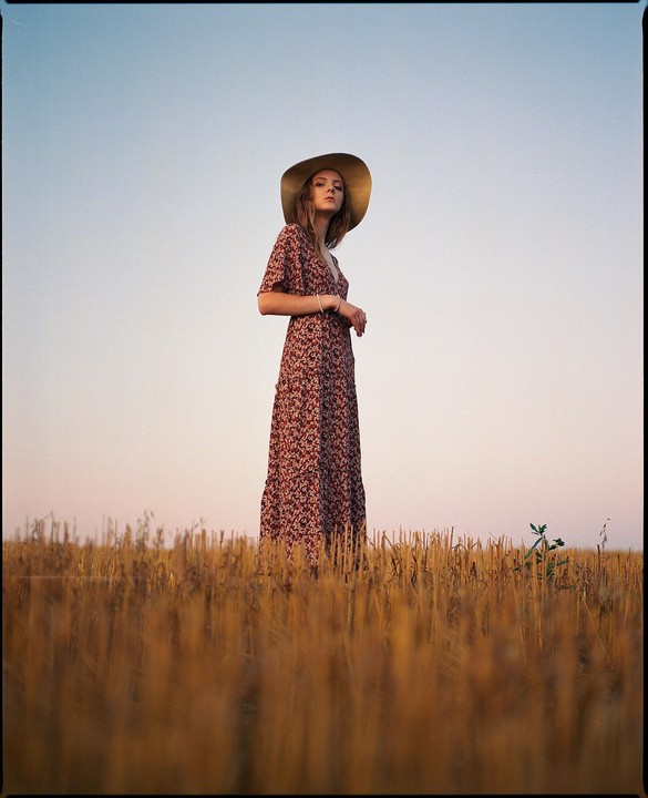 A woman in a floral dress and a wide-brimmed hat stands in a field of cut crops under a clear sky.