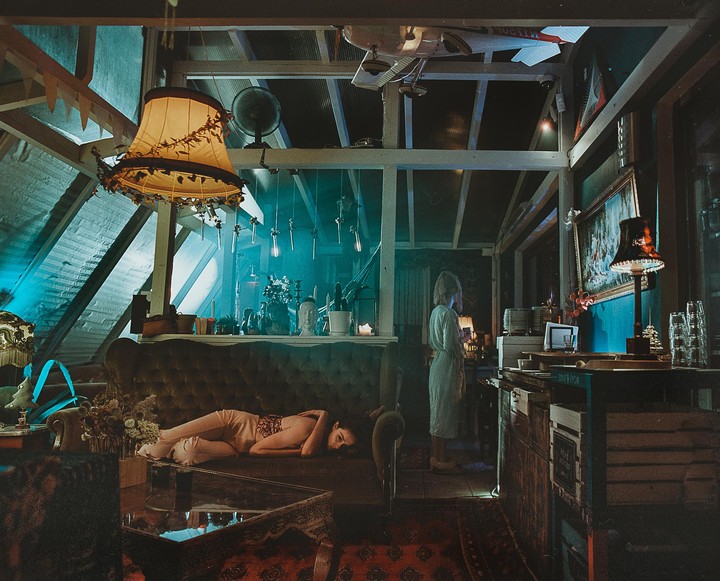 A woman lies on a sofa while another woman stands in a dimly lit, cluttered room with a sloped ceiling.
