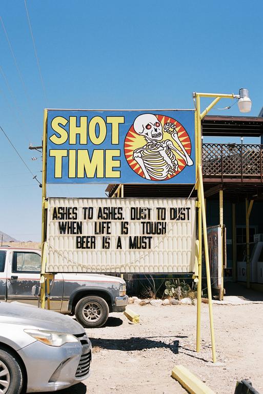 A sign with a skeleton illustration and text about "Shot Time" and "Beer is a Must" stands next to parked vehicles under a clear sky.