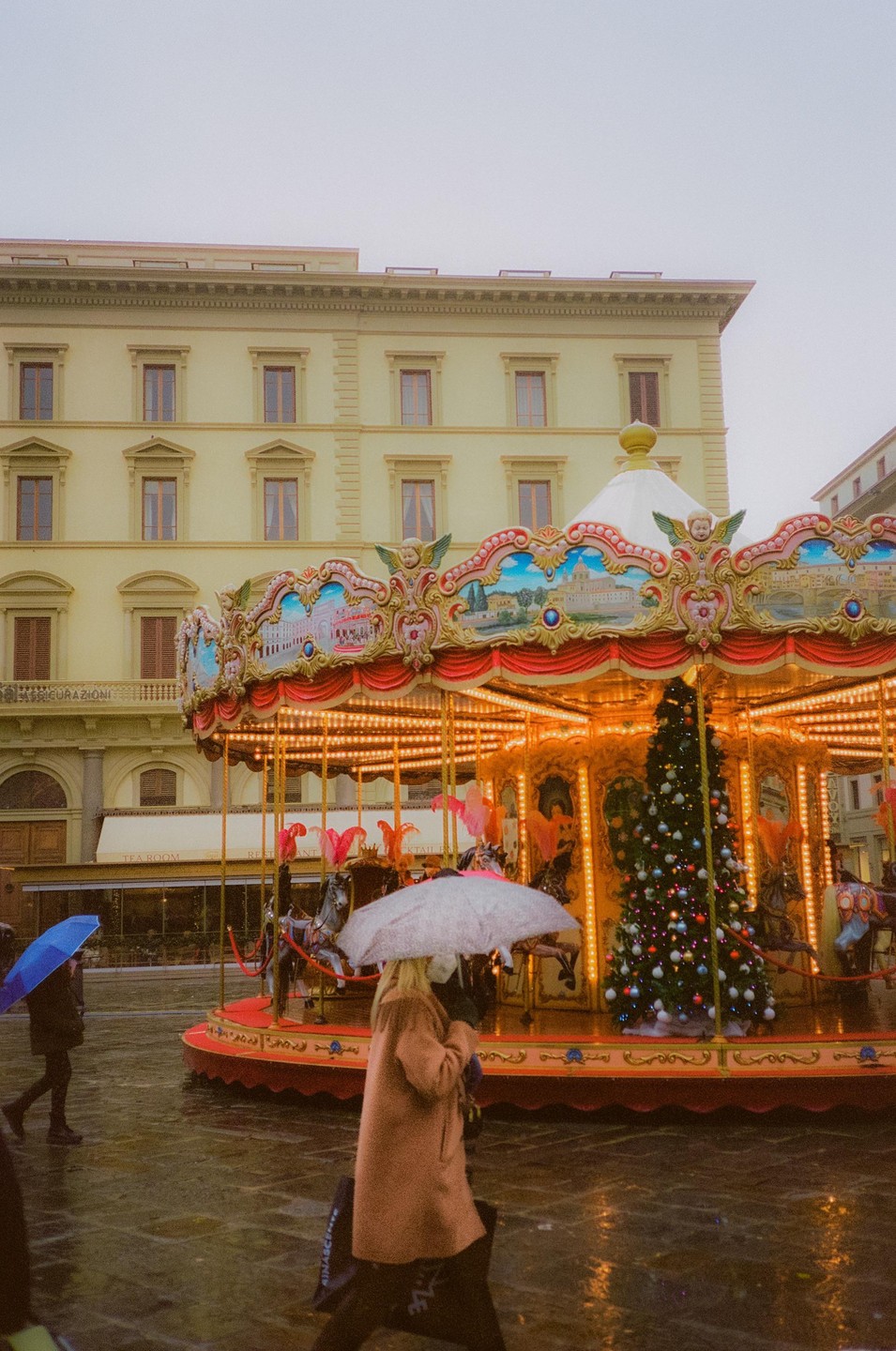 A woman with an umbrella walks past a brightly lit carousel with a Christmas tree on a rainy evening.