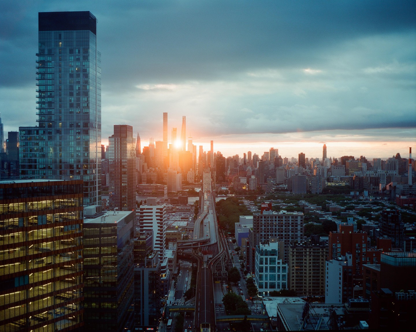 The sun sets behind a cityscape with a tall building in the foreground and a bridge in the distance.