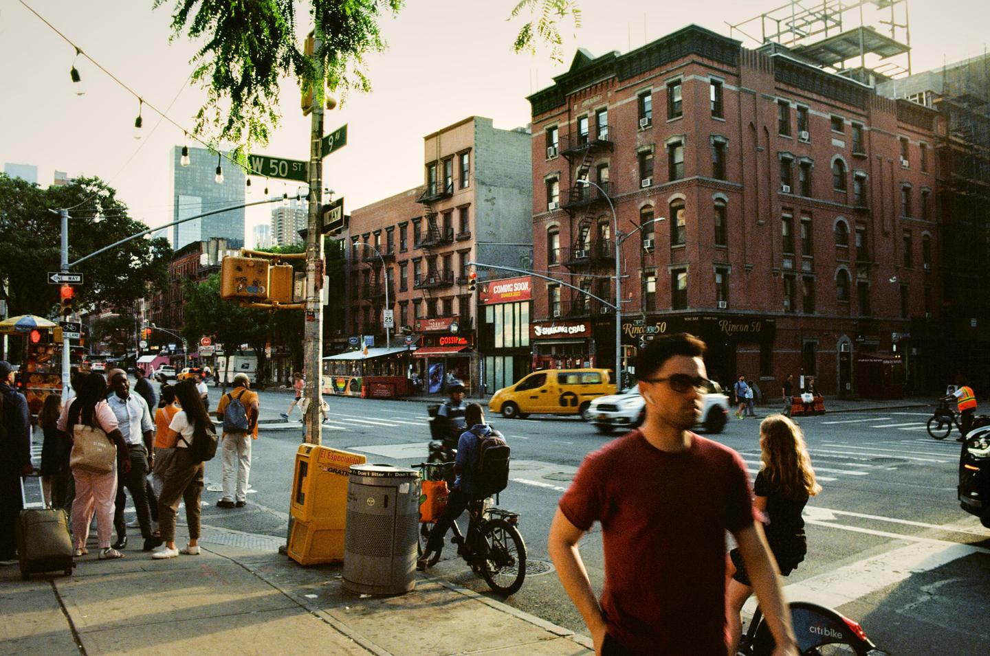 A street corner in New York City is bustling with people, vehicles, and brick buildings under a bright sky.