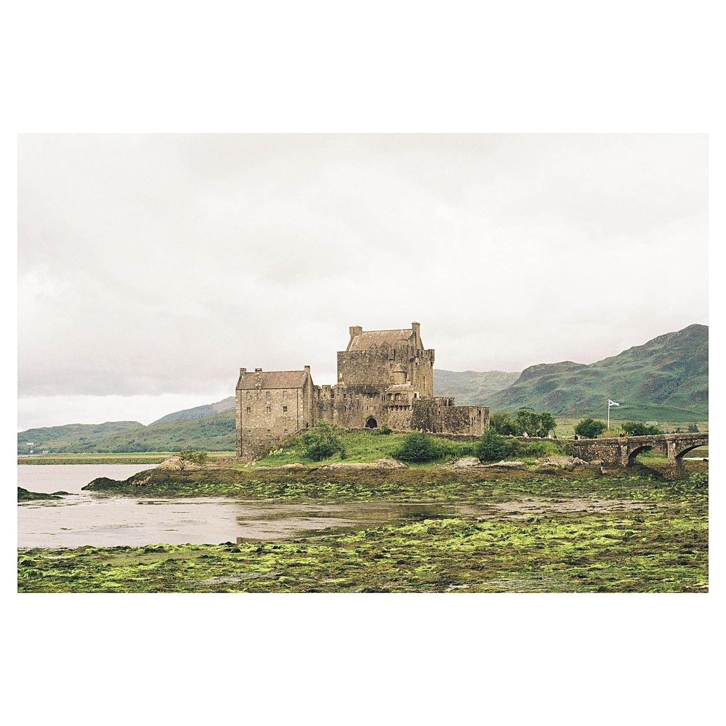 A stone castle stands on a small island connected by a bridge to a mountainous landscape under a cloudy sky.