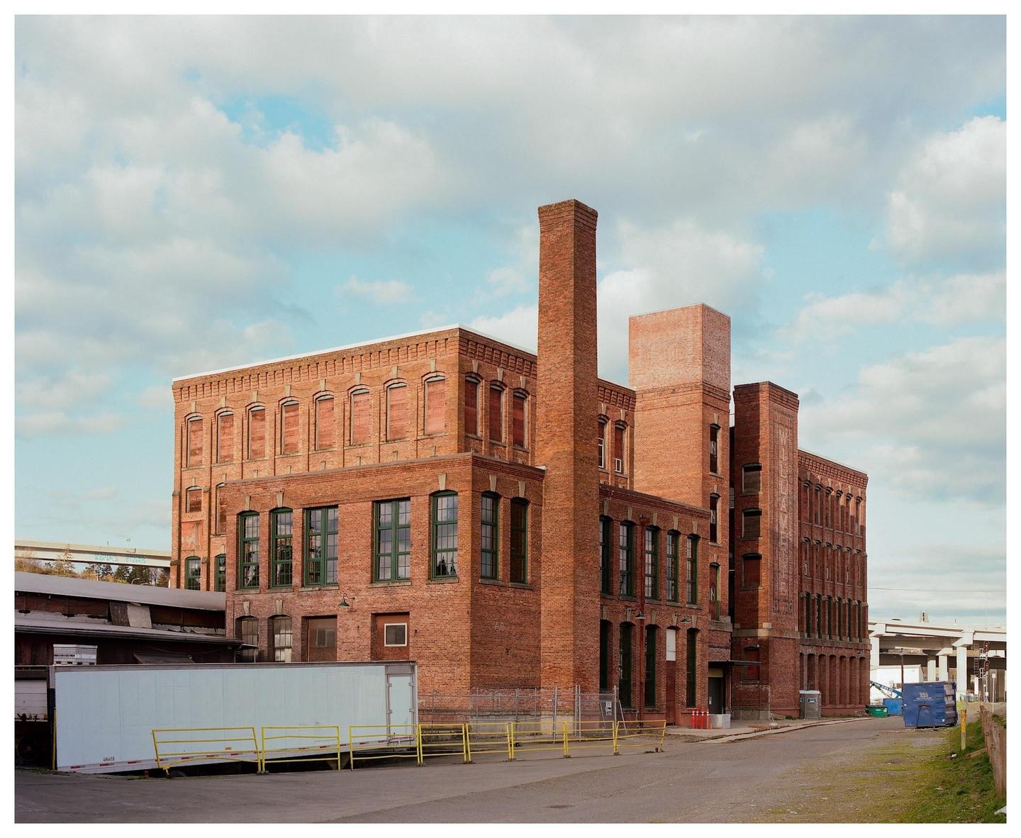 A large brick industrial building with a tall chimney stands under a partly cloudy sky.