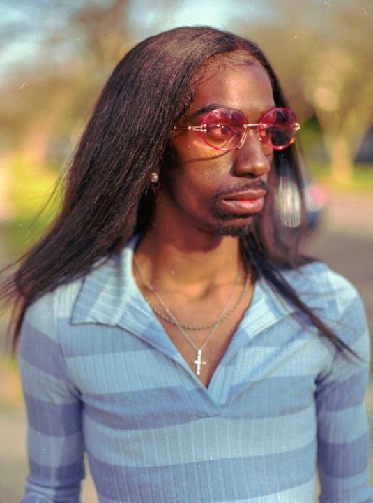 A person with long dark hair, a beard, and pink sunglasses wears a striped blue shirt and a cross necklace.