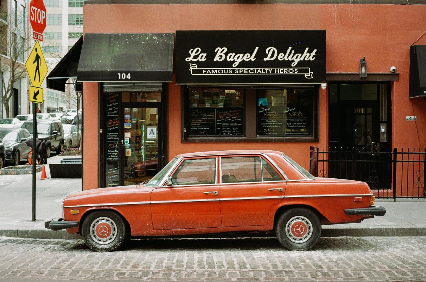 An orange car is parked on a cobblestone street in front of a bagel shop.