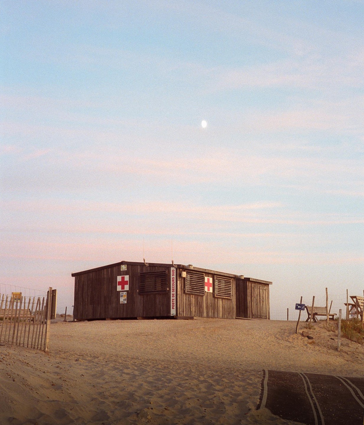 A wooden lifeguard hut with red crosses on its side sits on a sandy beach under a pale sky with a visible moon.