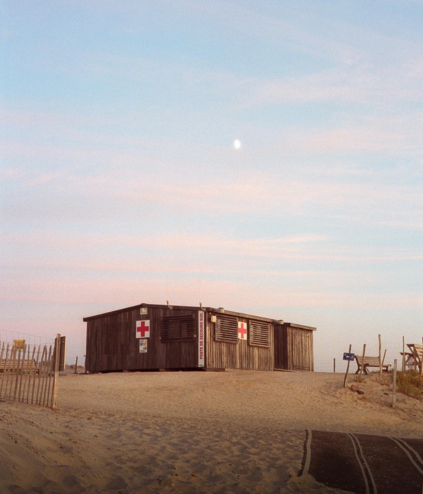 A wooden lifeguard hut with red crosses on its side sits on a sandy beach under a pale sky with a visible moon.
