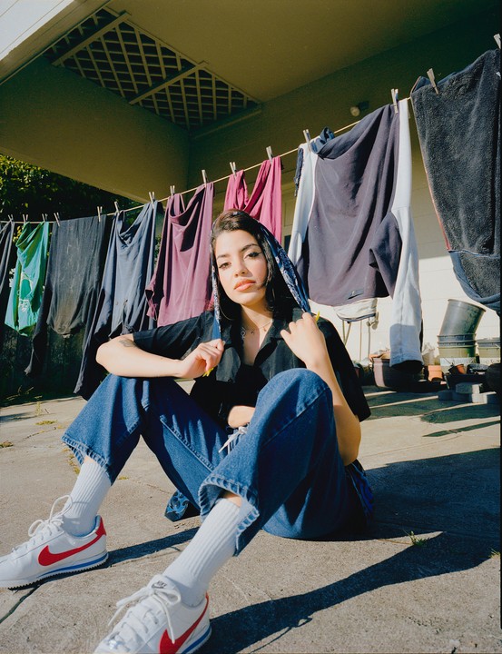 A woman sits on the ground with laundry hanging on a clothesline behind her.