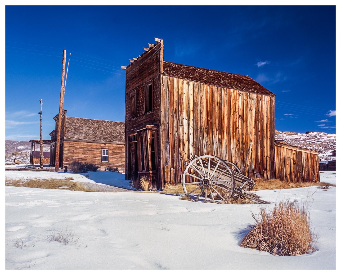 An old wooden building with a wagon wheel in front of it stands in a snow-covered landscape under a bright blue sky.
