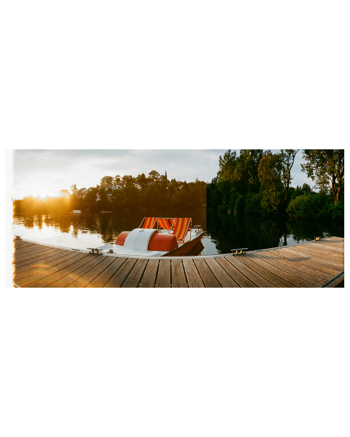 A pedal boat with striped seats is docked at a wooden pier on a lake during sunset, with a town and trees on the far bank.