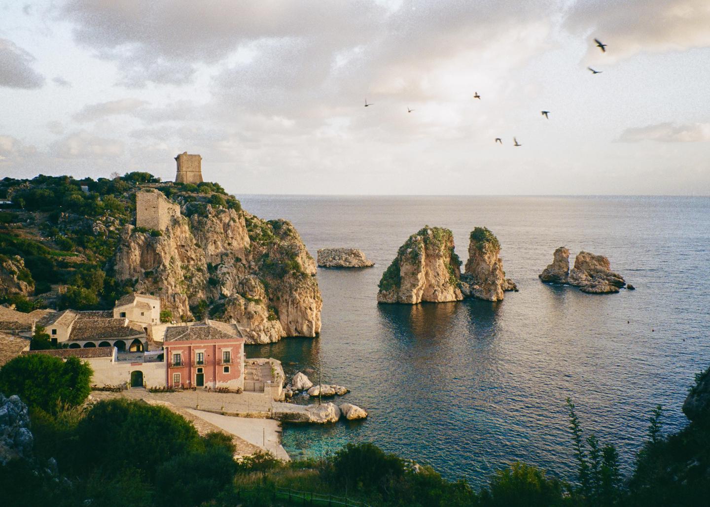 A small village with a pink building and a tower on a cliff overlooks the sea with several rock formations and birds flying in the sky.