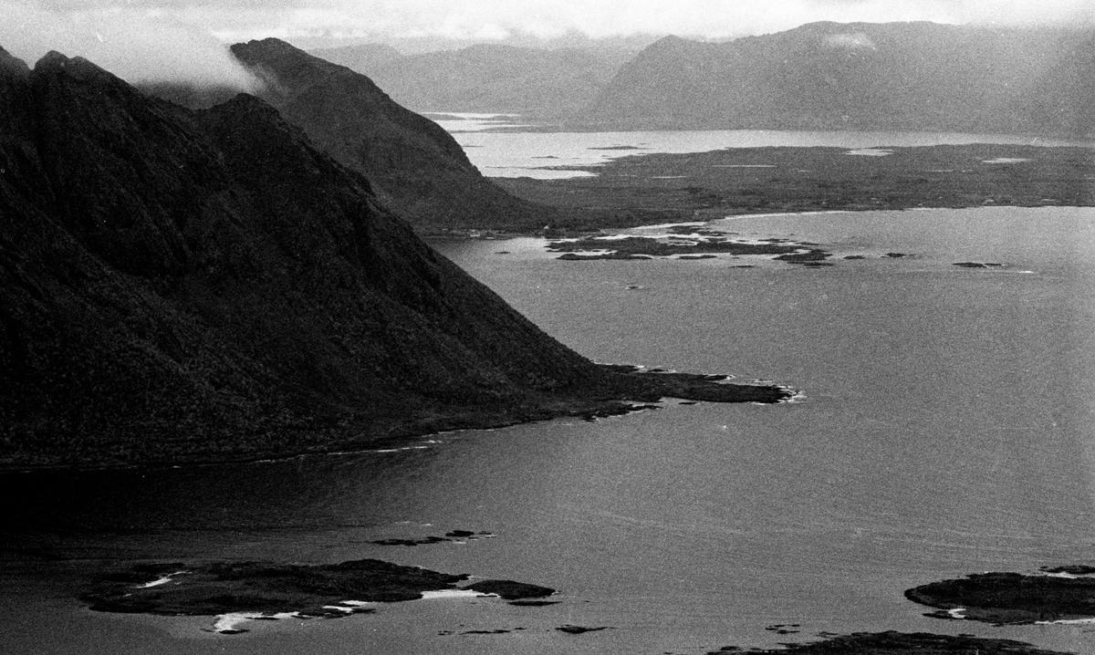 A black and white aerial landscape shows a body of water with small islands and a mountainous coastline under a cloudy sky.