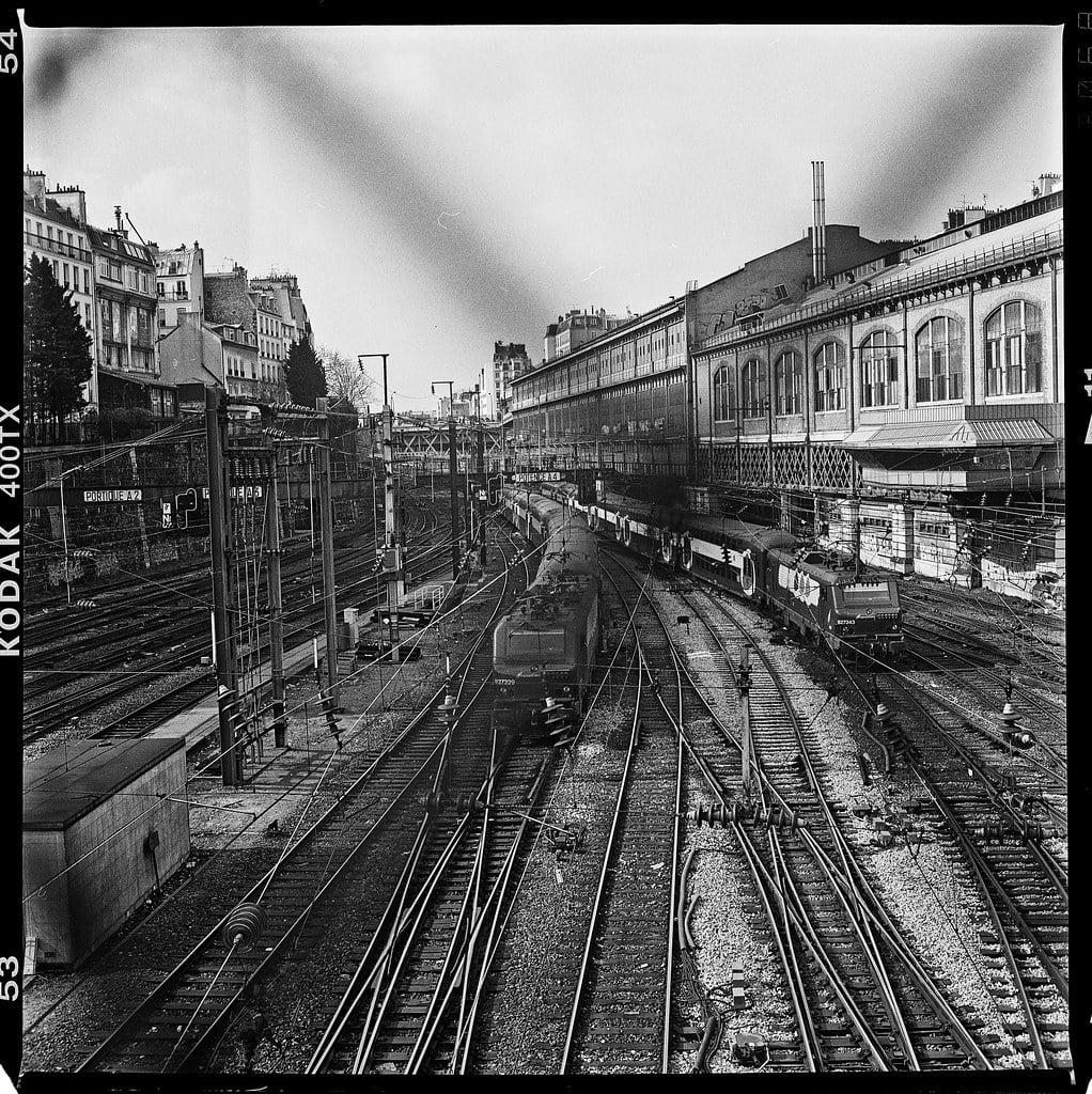 A black and white high-angle shot shows a train station with multiple tracks and trains, with buildings on either side.