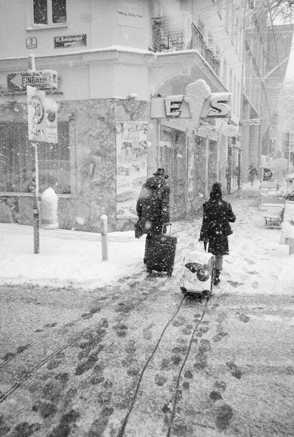 Two people pull luggage through a snowy street past a building with an "EIS" sign.