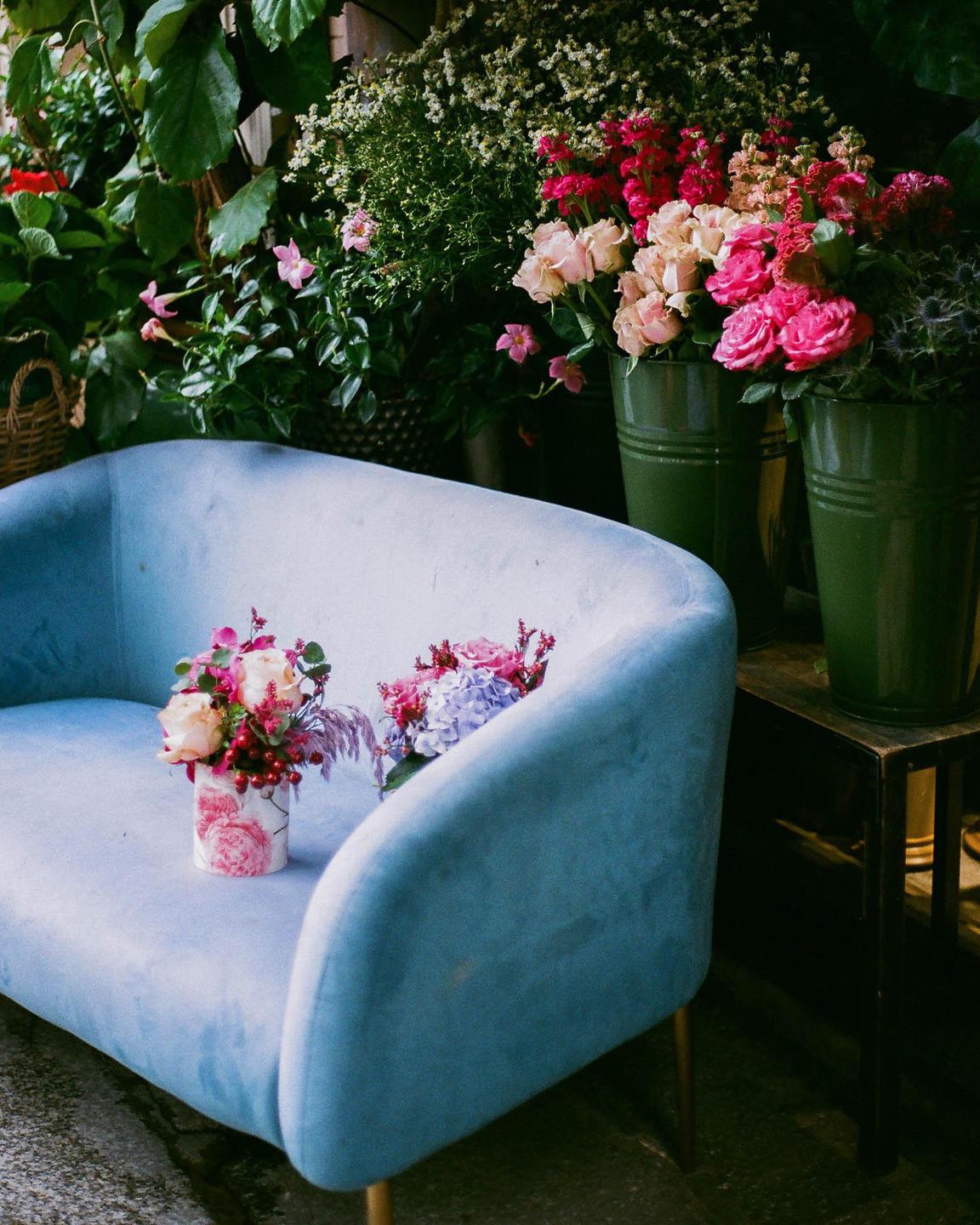 Two small bouquets sit on a light blue sofa in front of a display of flowers and plants.