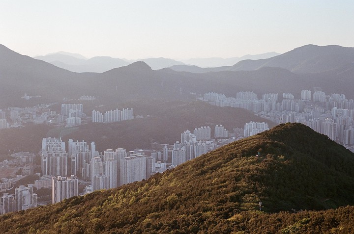 A city with many tall buildings is nestled among several layers of mountains under a hazy sky.