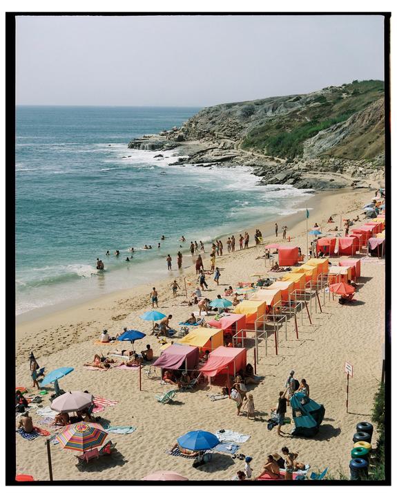 A crowded beach with colorful umbrellas and tents is visible next to a rocky coastline and the ocean.
