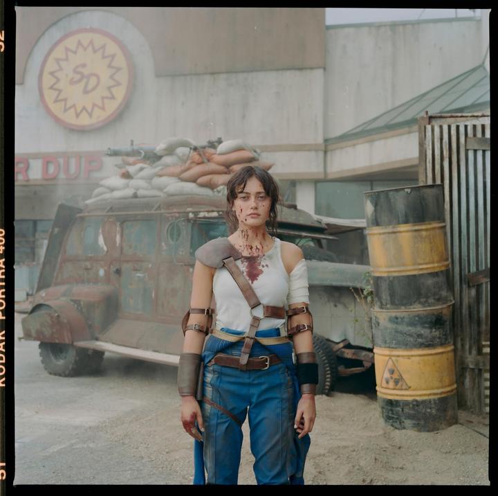 A woman with blood on her face and shirt stands in front of a rusty vehicle and barrels.