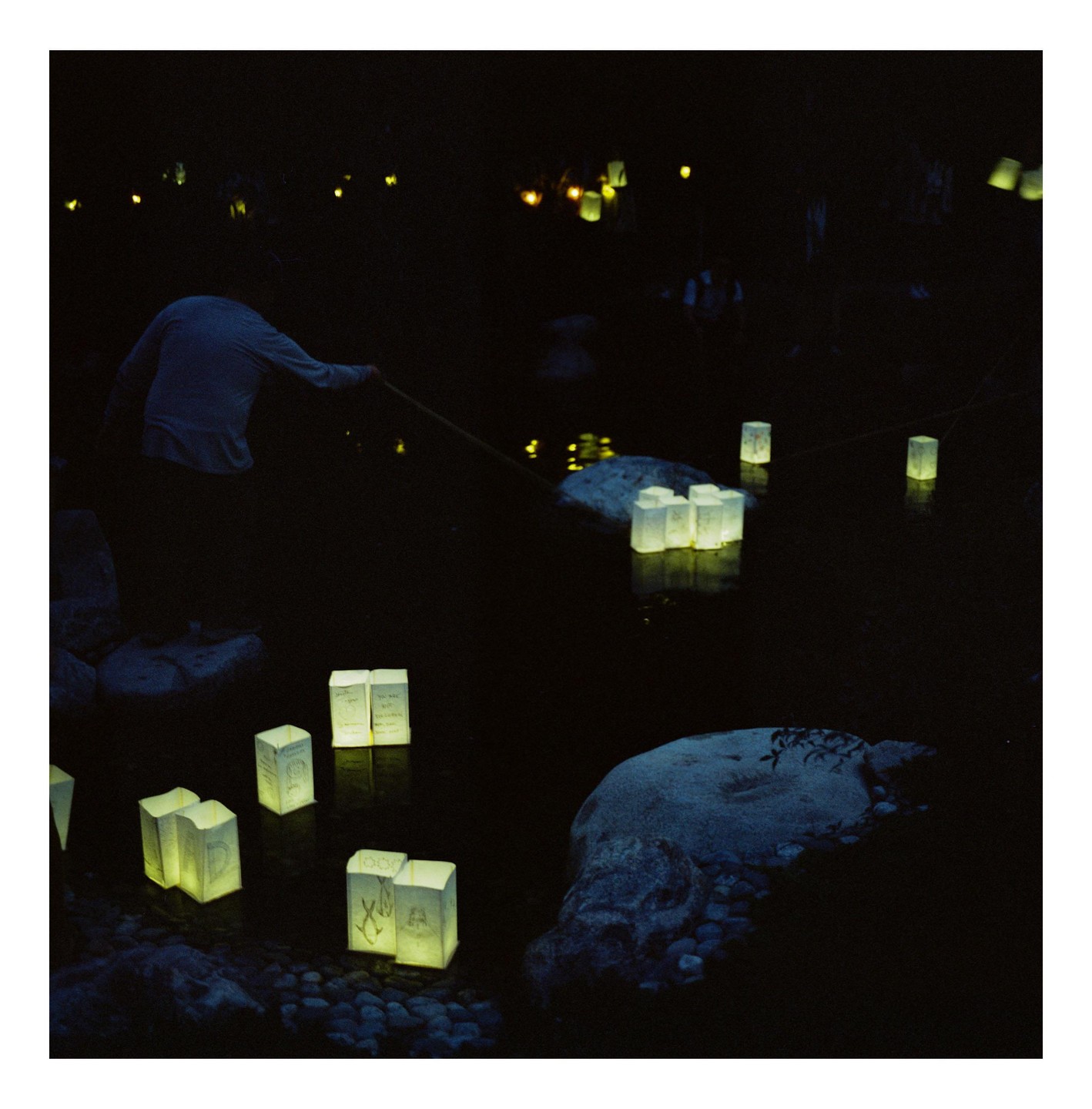 A person pushes a floating lantern with a stick in a dark body of water surrounded by rocks and other lanterns.