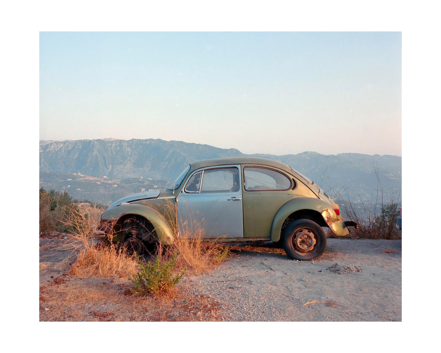 A two-tone Volkswagen Beetle is parked on a dirt road overlooking a valley and mountains.