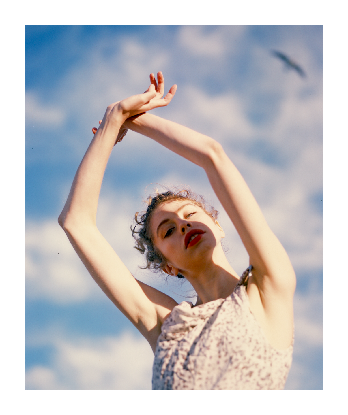 A woman with red lipstick and her arms raised is looking up at the camera against a blue sky with clouds.