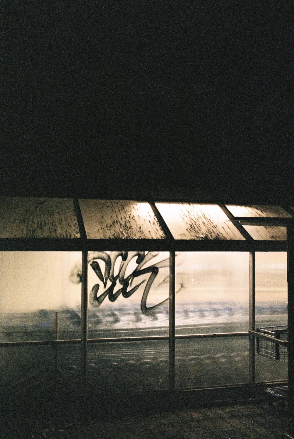 A glass shelter with graffiti on its window stands under a dark sky at night.