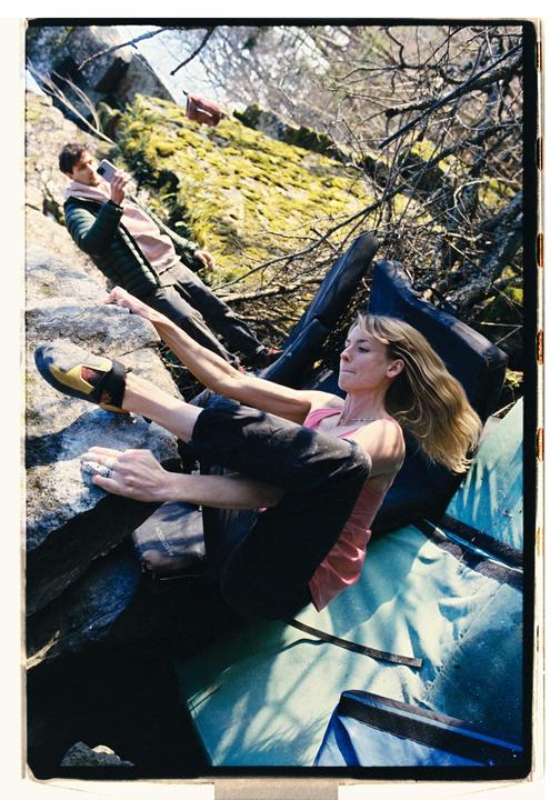 A woman bouldering on a rock with a man filming her in the background.