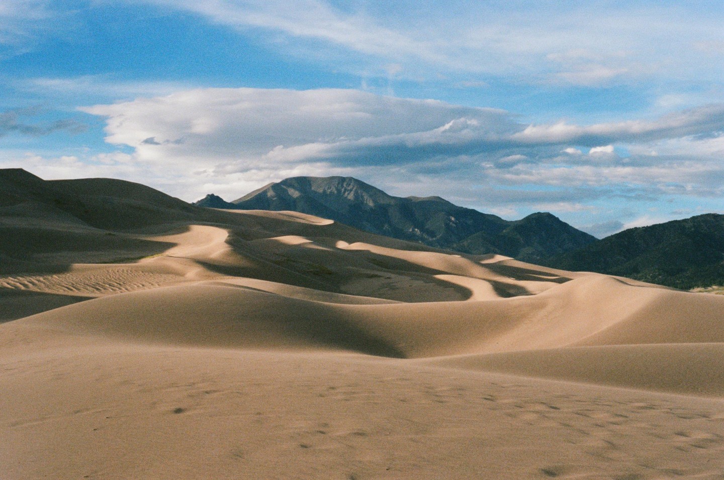 A vast desert landscape features sand dunes in the foreground and mountains under a cloudy sky in the background.