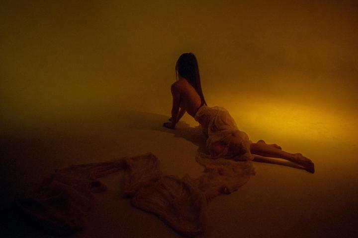 A woman with long hair is on her hands and knees, looking away from the camera, with a long piece of fabric draped around her lower body.