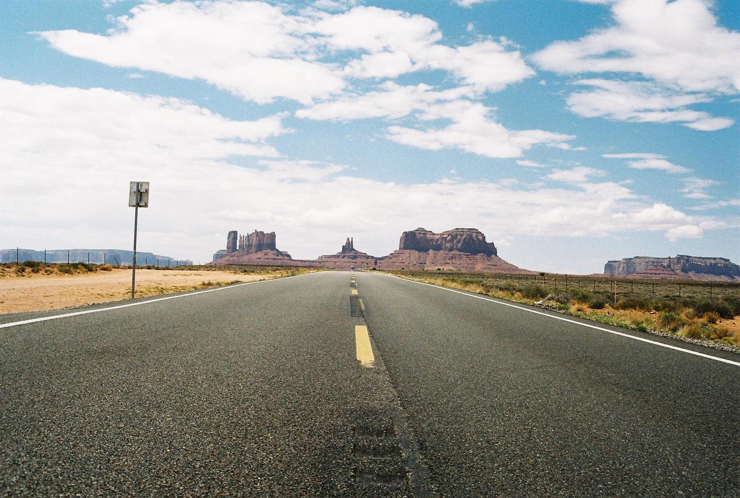 A long, straight road leads toward distant mesas under a sky with scattered clouds.