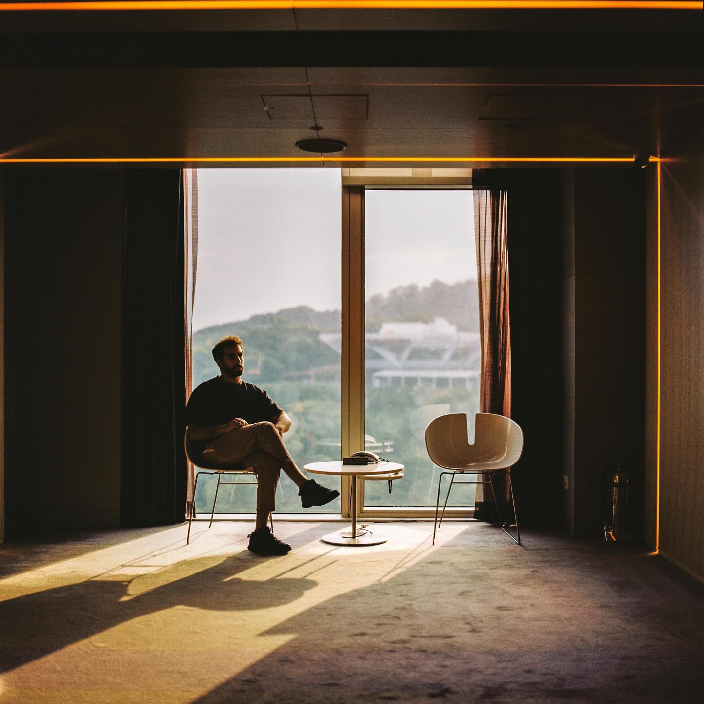 A man sits in a chair by a large window, with sunlight streaming into the room.