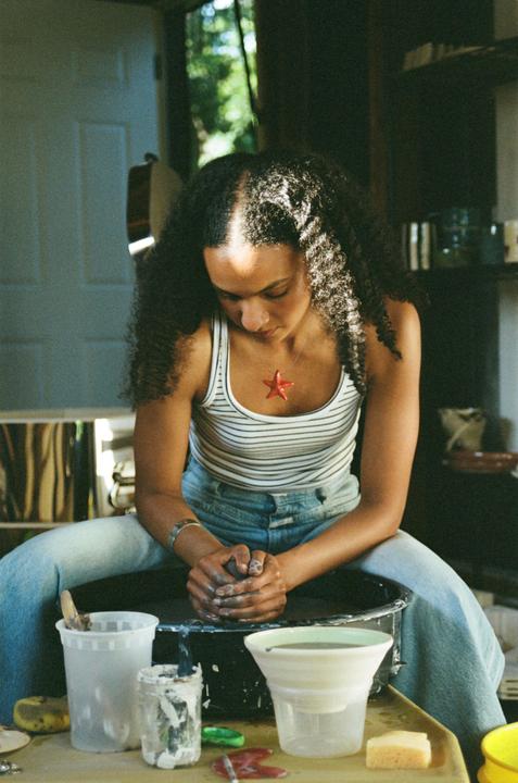 A woman with curly hair is seated at a pottery wheel, looking down at her hands.