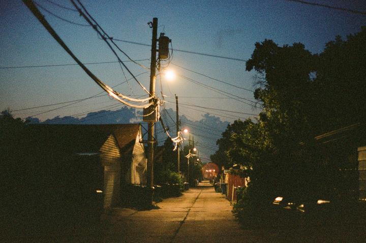 A narrow alleyway is illuminated by streetlights at night, with houses and trees lining its sides.