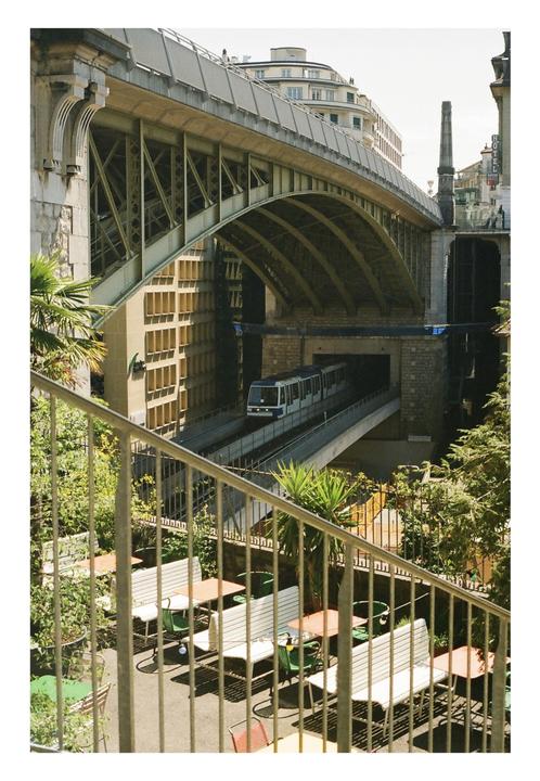 A train emerges from a tunnel under a large bridge, with an outdoor cafe visible in the foreground.