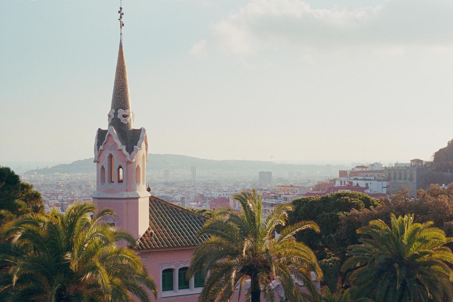 A pink turret with a pointed roof stands above palm trees and a city.