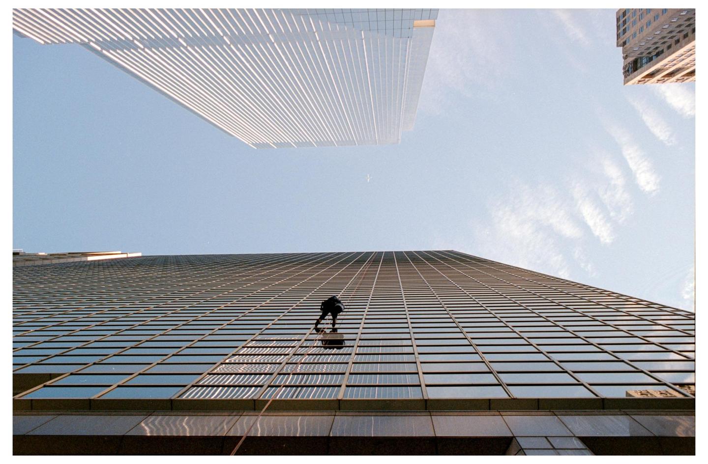 A window washer dangles from ropes on the side of a tall glass building, with another skyscraper visible in the background against a blue sky.