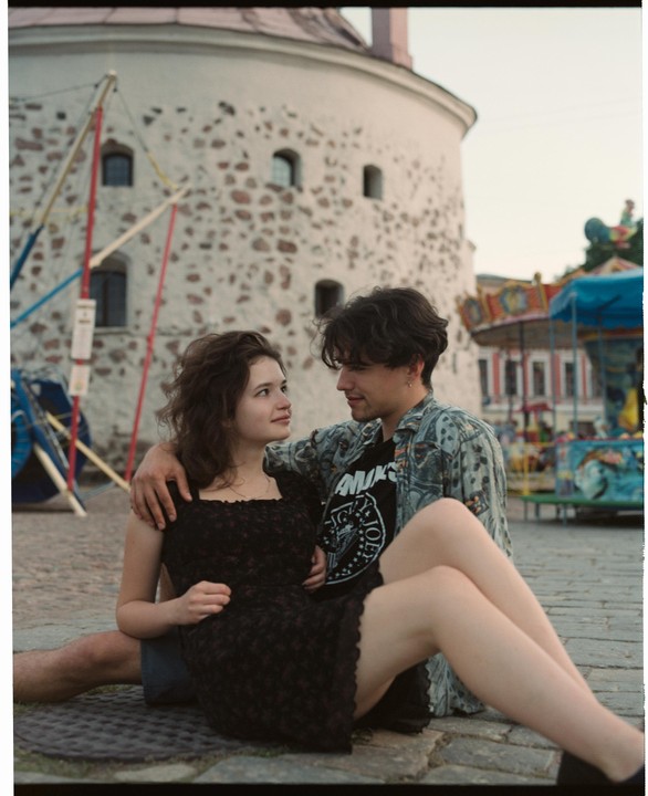 A young couple sits on the ground in front of a stone building and a carousel.