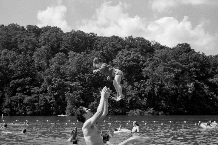 A man throws a child into the air in a lake with a forested hill in the background.