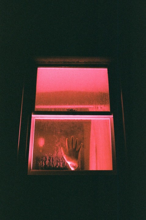A hand is pressed against a steamy window, illuminated by a red light from inside.