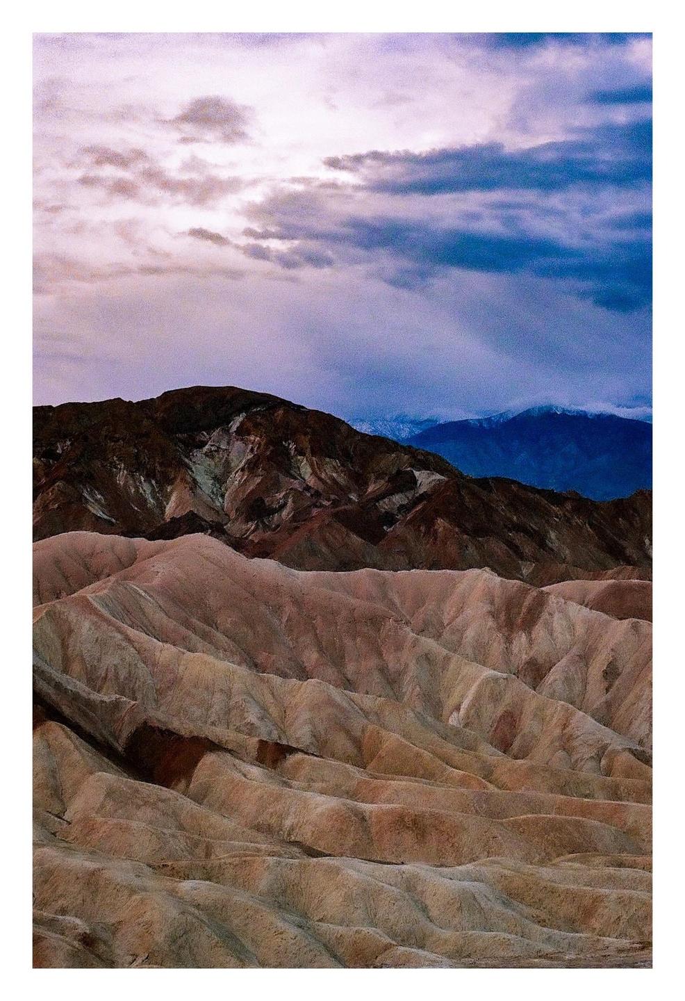 A dramatic sky with purple and blue clouds hangs over a desert landscape with rolling hills and mountains.
