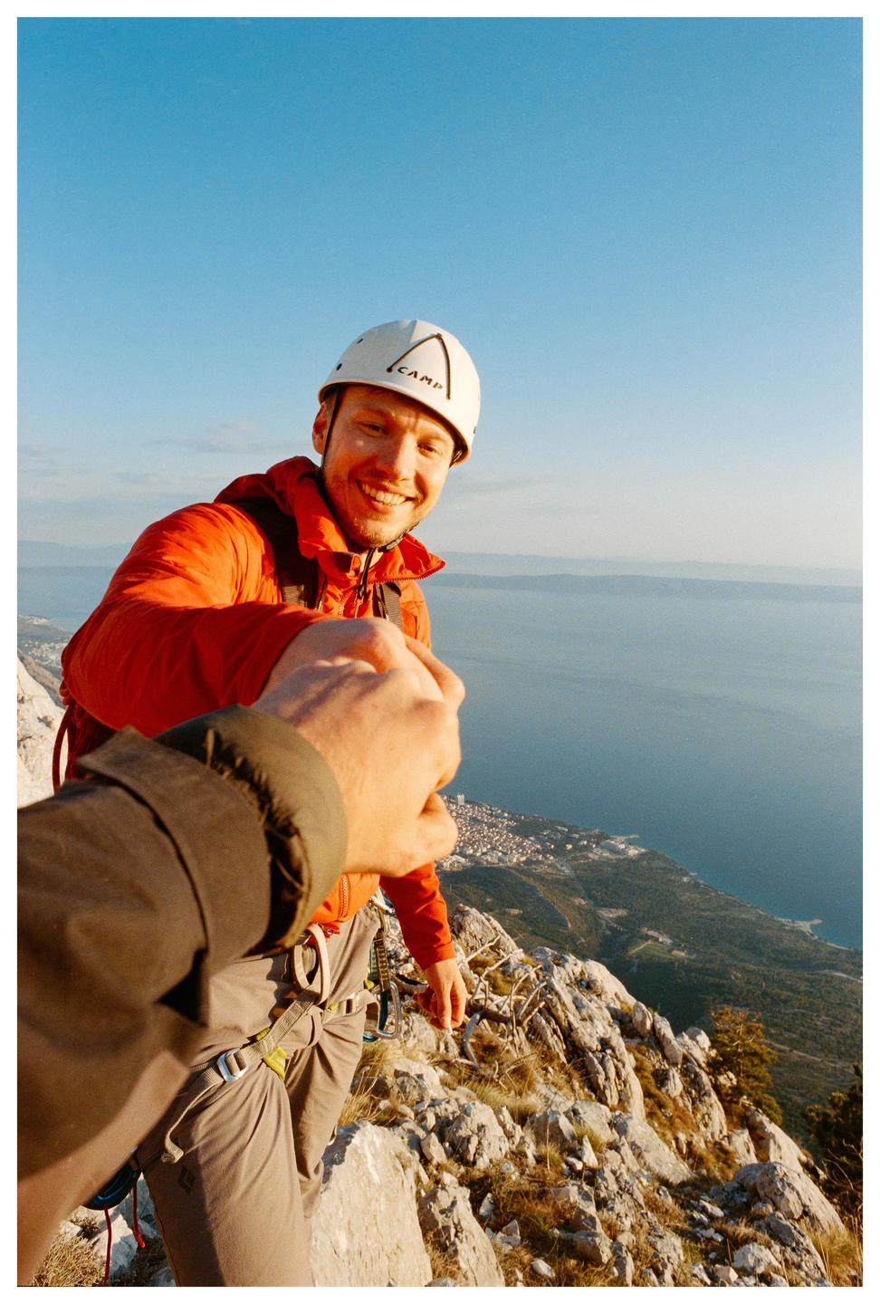 A man in a climbing helmet and orange jacket smiles at the camera while standing on a rocky mountain overlooking a town and the sea.