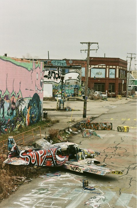 A small airplane covered in graffiti sits on a paved lot next to a building with a large pink mural.