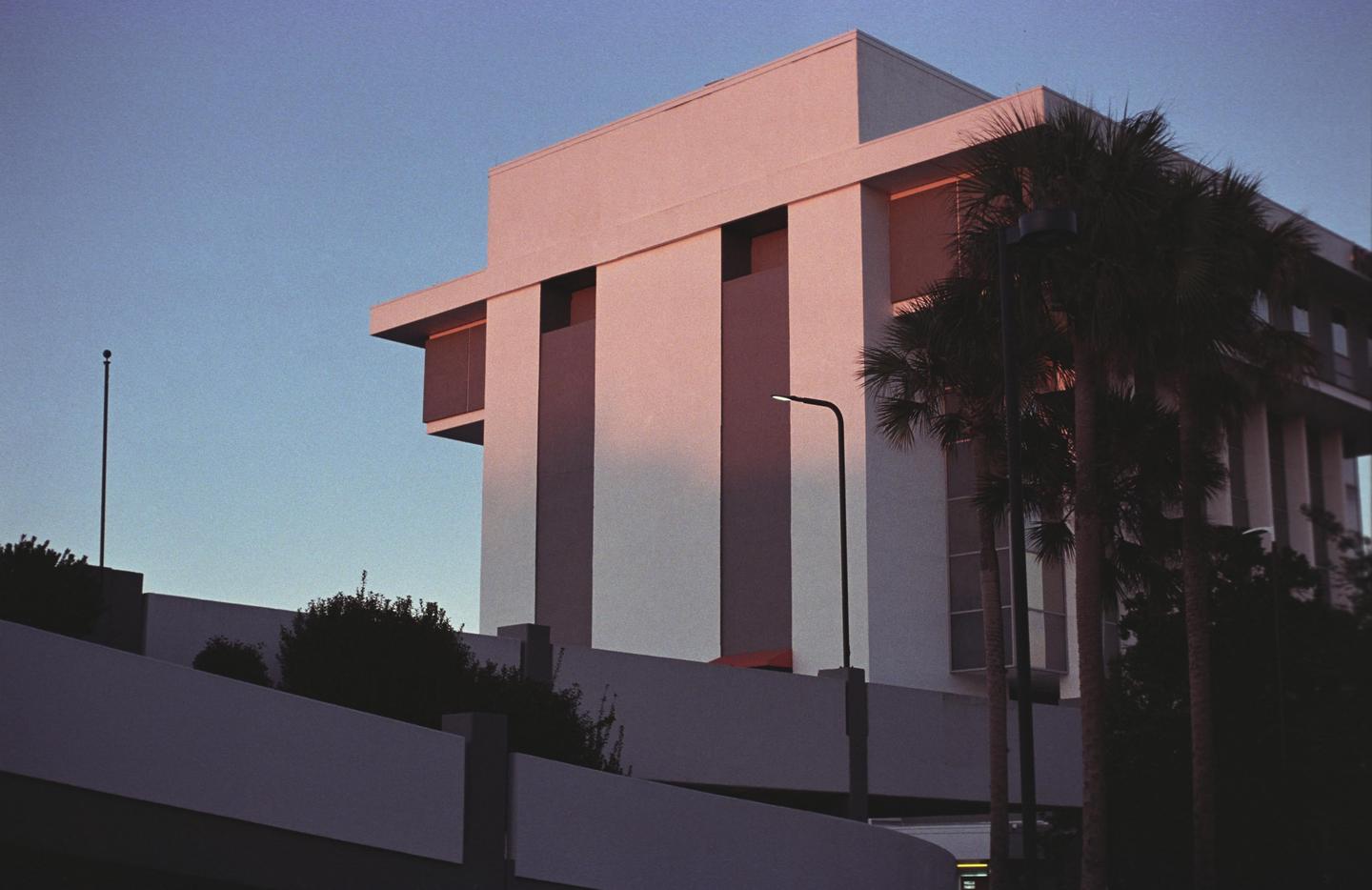 A modern building with palm trees and a street light is illuminated by the golden hour sun.