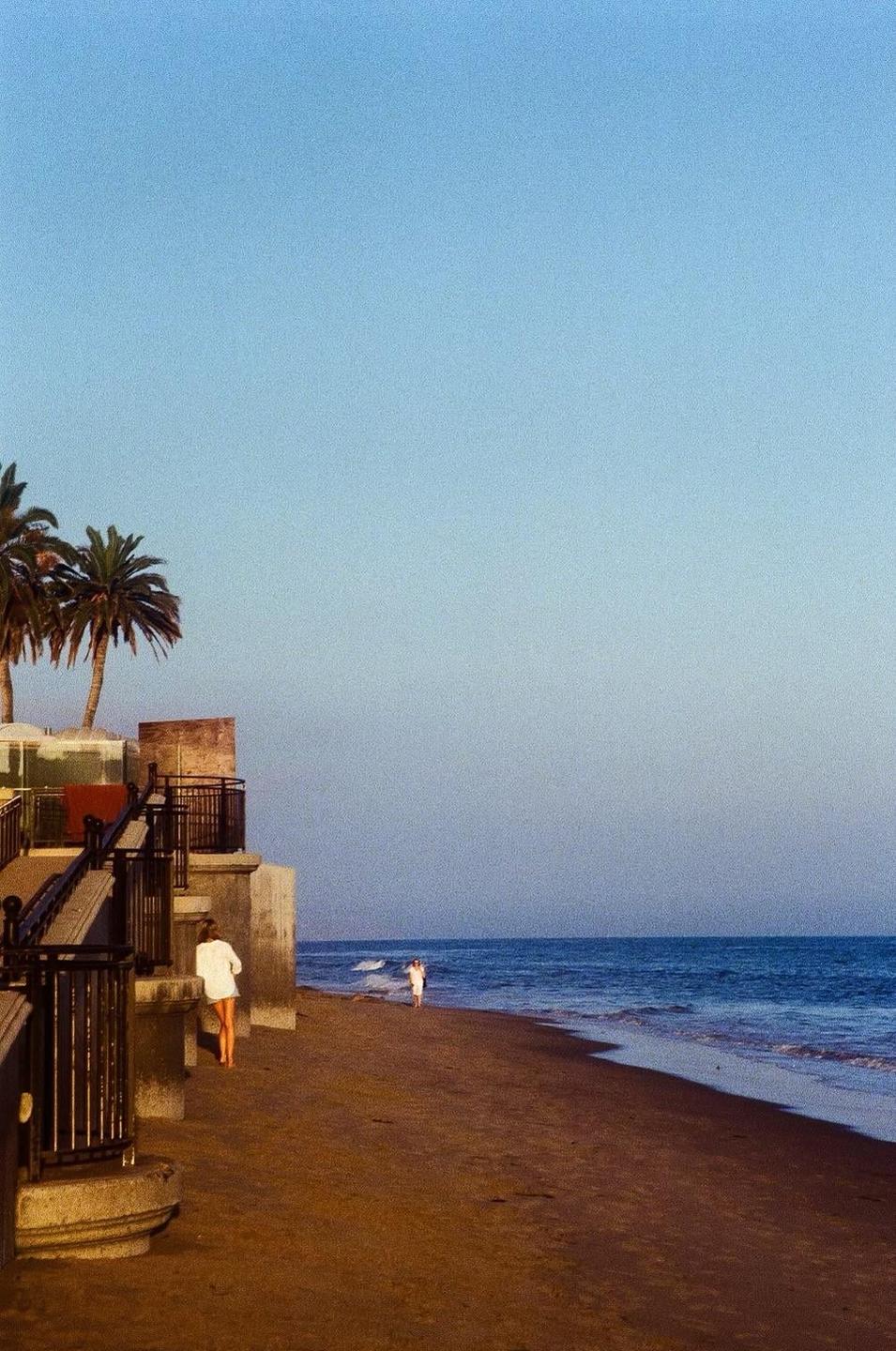 A woman walks on a sandy beach next to the ocean with palm trees and a building on the left.